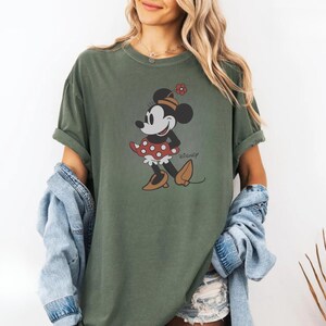 May include: Olive green t-shirt featuring a graphic of Minnie Mouse in a red polka dot dress and brown shoes. The word "Disney" is printed in a script font. The shirt has short sleeves and a relaxed fit.