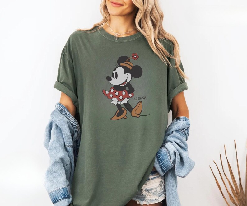 May include: Olive green t-shirt featuring a graphic of Minnie Mouse in a red polka dot dress and brown shoes. The word "Disney" is printed in a script font. The shirt has short sleeves and a relaxed fit.