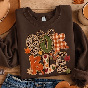 May include: A dark chocolate brown crewneck sweatshirt featuring the text GOBBLE in large, stylized letters. The letters are decorated with leopard print, orange plaid, and autumn leaf patterns, accented by a leopard print bow and a small turkey.