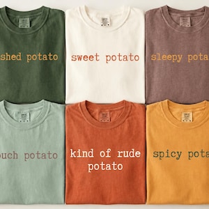 Op de afbeelding: Zes t-shirts met ronde hals in verschillende kleuren, waaronder olijfgroen, crème, mauve, saliegroen, terracotta en mosterdgeel. Elk shirt heeft tekst op de borst gedrukt: mashed potato, sweet potato, sleepy potato, couch potato, kind of rude potato en spicy potato.