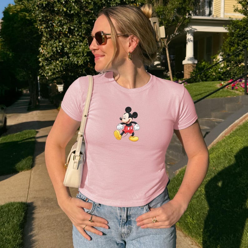 Puede incluir: Una camiseta de manga corta en color rosa claro con un gr&aacute;fico central de Mickey Mouse con sus cl&aacute;sicos pantalones cortos rojos, zapatos amarillos y guantes blancos. La prenda se combina con vaqueros azules y un peque&ntilde;o bolso de hombro color crema.