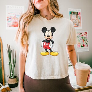 May include: A cream-colored short-sleeve t-shirt featuring a centered graphic of Mickey Mouse. The character is depicted with his signature black ears, red shorts, white gloves, and yellow shoes, standing with hands on his hips against the fabric.