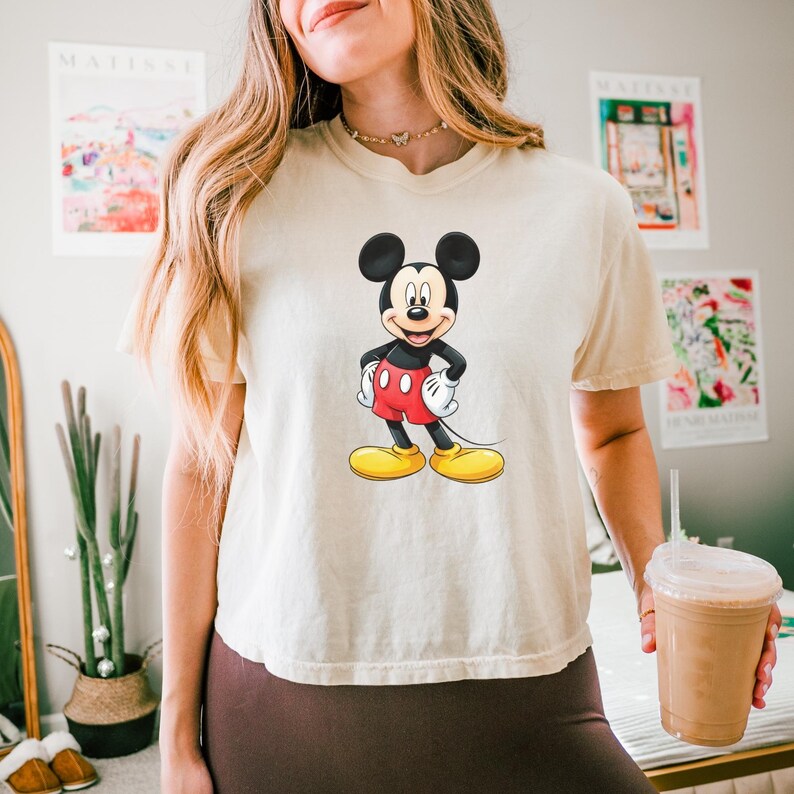 May include: A cream-colored short-sleeve t-shirt featuring a centered graphic of Mickey Mouse. The character is depicted with his signature black ears, red shorts, white gloves, and yellow shoes, standing with hands on his hips against the fabric.