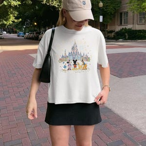 K&ouml;nnte beinhalten: Wei&szlig;es T-Shirt mit einer farbenfrohen Illustration eines Schlosses und Zeichentrickfiguren. Die Grafik zeigt Micky Maus, Minnie Maus, Donald Duck, Goofy und andere Charaktere. Das Shirt wird mit einem schwarzen Rock kombiniert.