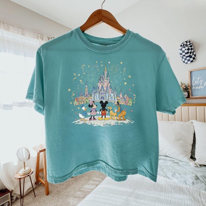 K&ouml;nnte beinhalten: T&uuml;rkisfarbenes T-Shirt mit einem farbenfrohen Aufdruck von Disney-Figuren, Mickey Mouse, Minnie Mouse, Donald Duck und Pluto, vor einem Schloss. Die Grafik enth&auml;lt Feuerwerk und Konfetti. Das Shirt h&auml;ngt an einem Holzb&uuml;gel.