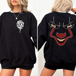 May include: A black long-sleeve sweatshirt featuring a white graphic on the front left chest that reads "YOU'LL FLOAT TOO" in the shape of a balloon. The back displays a large, detailed graphic of a clown face with red and dark features.