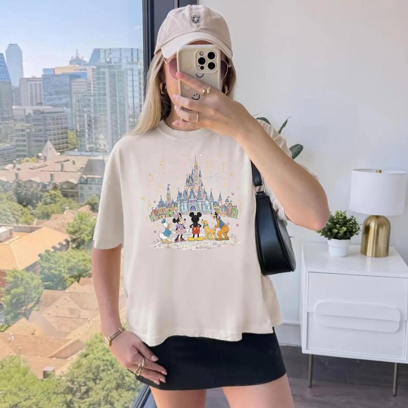 K&ouml;nnte beinhalten: Cremefarbenes T-Shirt mit einem farbenfrohen Aufdruck von Disney-Figuren vor einem Schloss. Das Shirt hat kurze &Auml;rmel und einen Rundhalsausschnitt. Die Person tr&auml;gt eine beige Kappe und einen schwarzen Rock.