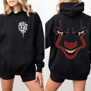 May include: A black hooded sweatshirt featuring a white balloon graphic on the front chest with the text "YOU'LL FLOAT TOO". The back of the hoodie displays a large, detailed graphic of a clown face with red makeup, yellow eyes, and a wide grin.