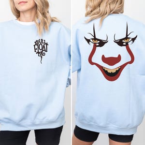 May include: A light blue long-sleeve sweatshirt featuring a black graphic of a balloon with the text "YOU'LL FLOAT TOO" on the front left chest and a large, colorful clown face graphic printed on the back, showcasing red, black, and yellow details.