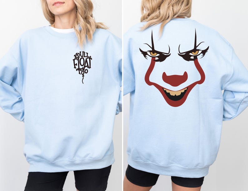 May include: A light blue long-sleeve sweatshirt featuring a black graphic of a balloon with the text "YOU'LL FLOAT TOO" on the front left chest and a large, colorful clown face graphic printed on the back, showcasing red, black, and yellow details.