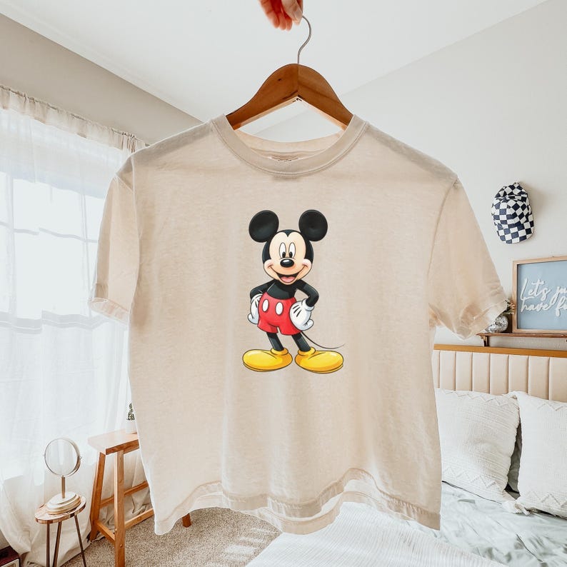 May include: A beige short-sleeved t-shirt featuring a centered graphic of Mickey Mouse. The character is depicted with his signature black ears, red shorts, white gloves, and yellow shoes, standing with hands on his hips against the light fabric.