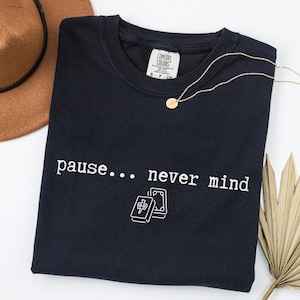 May include: A black short-sleeve t-shirt with a white graphic print that reads "pause... never mind" above an illustration of two mahjong tiles. The shirt features a Comfort Colors brand label at the inner neckline and a crew neck collar design.