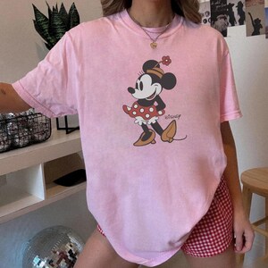 May include: A light pink t-shirt featuring a Minnie Mouse graphic. The cartoon character is depicted in a red polka-dot dress, brown shoes, and a matching hat with a flower. The Disney logo is printed below the character.
