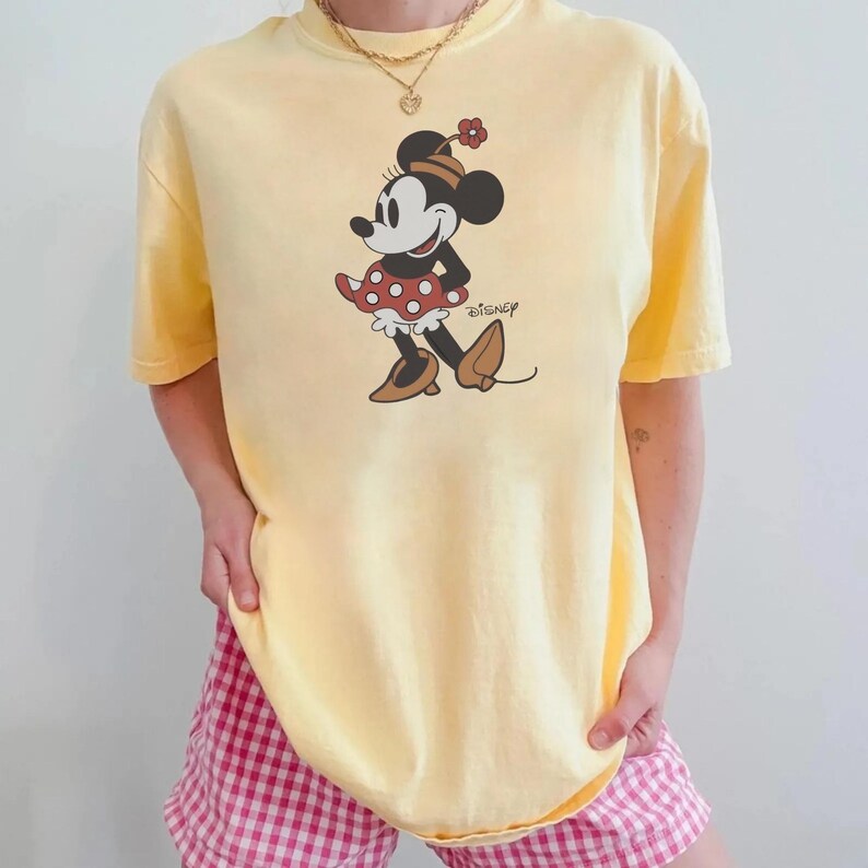 May include: A yellow t-shirt featuring a graphic of Minnie Mouse in a red polka dot dress, brown shoes, and a flower on her head. The Disney logo is printed below the character. The shirt is paired with pink and white checkered shorts.