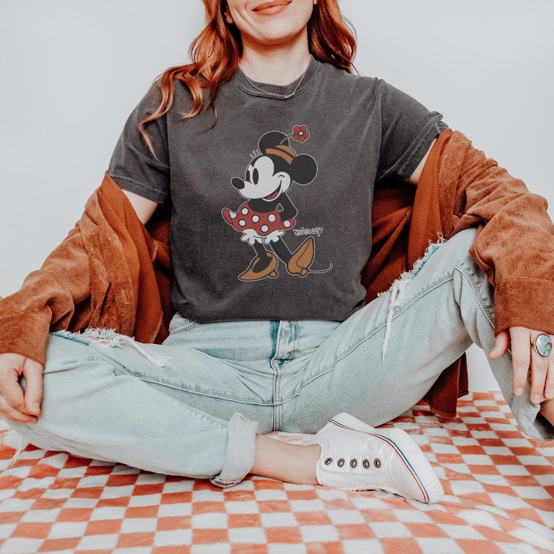 May include: Dark gray t-shirt featuring a vintage-style Minnie Mouse graphic. Minnie is depicted in a red polka-dot dress and matching bow. The shirt is paired with light-wash jeans and white sneakers, creating a casual, everyday look.