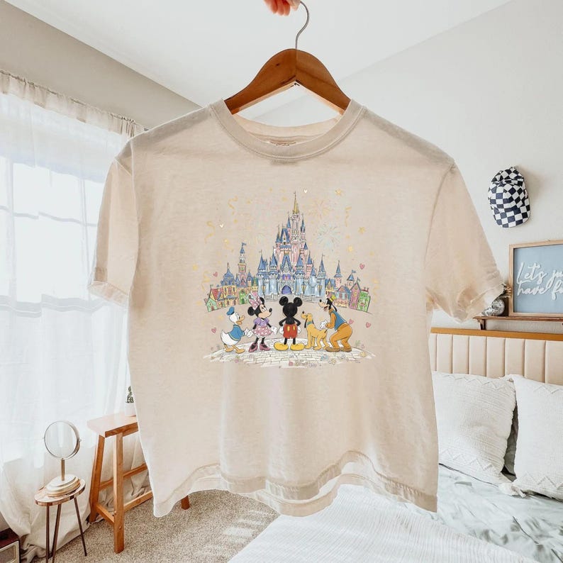 K&ouml;nnte beinhalten: Beiges T-Shirt mit farbenfroher Grafik von Disney-Figuren Mickey Mouse, Minnie Mouse, Donald Duck, Goofy und einem Schloss. Das T-Shirt h&auml;ngt an einem Holzb&uuml;gel. Das T-Shirt hat kurze &Auml;rmel und einen Rundhalsausschnitt.