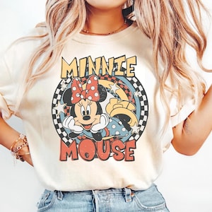 May include: Cream-colored t-shirt featuring a vintage-style graphic of Minnie Mouse. The design includes Minnie's face, a red polka-dot bow, and the words "MINNIE MOUSE" in a retro font. The graphic is set within a circular frame with a checkered pattern.