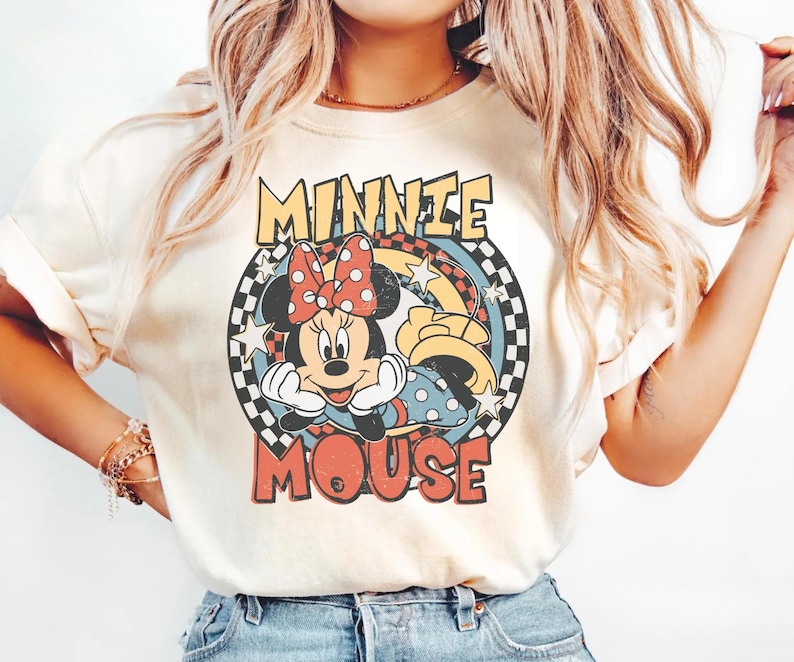 May include: Cream-colored t-shirt featuring a vintage-style graphic of Minnie Mouse. The design includes Minnie's face, a red polka-dot bow, and the words "MINNIE MOUSE" in a retro font. The graphic is set within a circular frame with a checkered pattern.