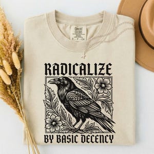 Puede incluir: Una camiseta beige con un estampado gráfico negro de un cuervo rodeado de elementos florales. El texto RADICALIZE está impreso sobre el ave y BY BASIC DECENCY debajo. La camiseta tiene cuello redondo y una etiqueta de Comfort Colors.