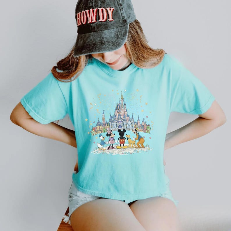 K&ouml;nnte beinhalten: Ein hellblaues T-Shirt mit einer farbenfrohen Grafik eines Schlosses und Zeichentrickfiguren. Zu den Figuren geh&ouml;ren Micky Maus, Minnie Maus, Donald Duck und Goofy. Die Person im Bild tr&auml;gt eine schwarze Kappe mit dem Wort "HOWDY".