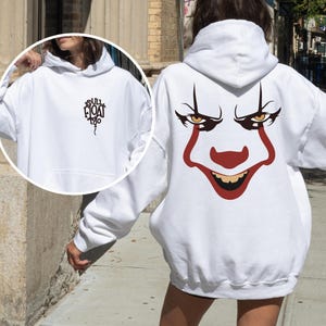May include: A white hooded sweatshirt featuring a large graphic of a clown face on the back with red and black details. The front chest area displays a small black graphic of a balloon with the text "YOU'LL FLOAT TOO" printed in a stylized font.