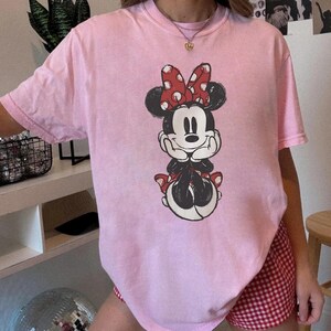 May include: A pale pink t-shirt with a graphic of Minnie Mouse. The cartoon character is in black and white with a red bow. The t-shirt is a relaxed fit, suitable for casual wear.