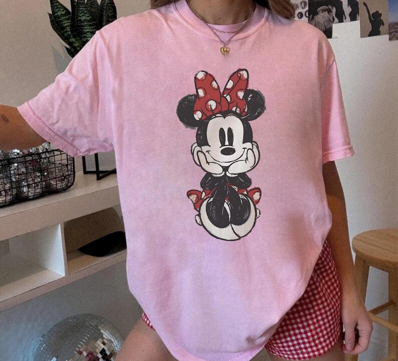 May include: A pale pink t-shirt with a graphic of Minnie Mouse. The cartoon character is in black and white with a red bow. The t-shirt is a relaxed fit, suitable for casual wear.