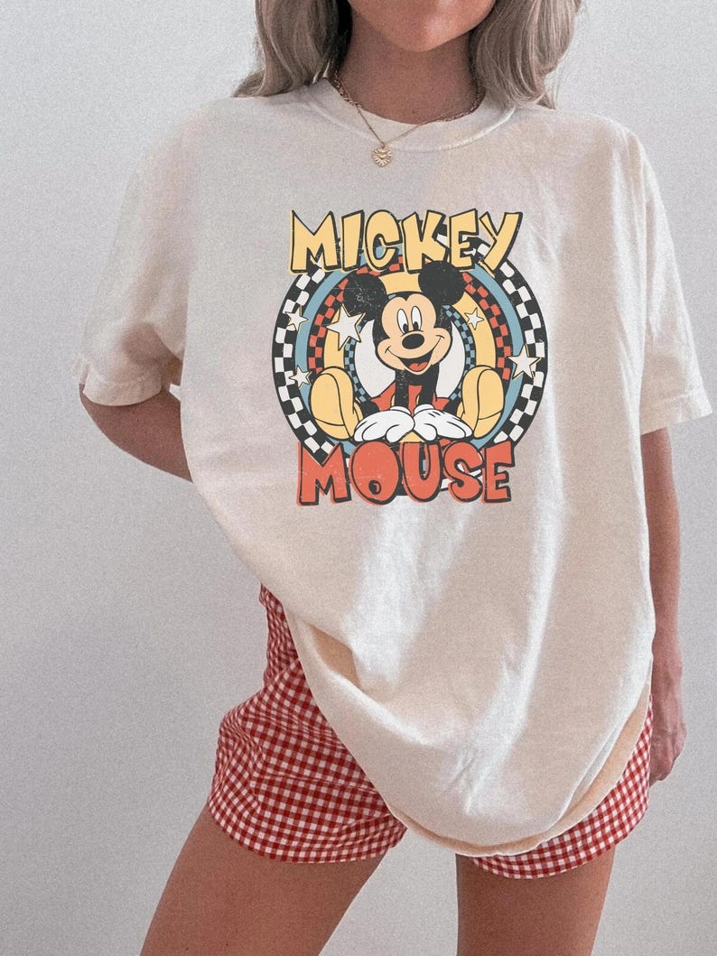 May include: Cream-colored t-shirt featuring a graphic of Mickey Mouse. The design includes the words "Mickey Mouse" in a retro font, set against a checkered and rainbow background. The shirt is paired with red and white checkered shorts.