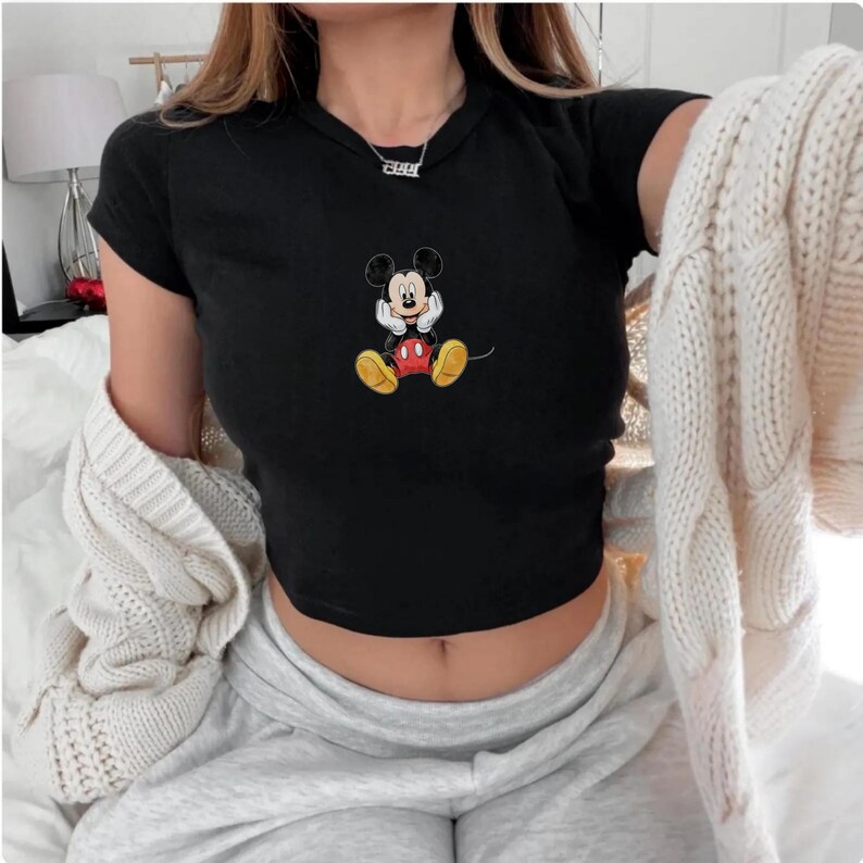 May include: Black crop top with a colourful embroidered Mickey Mouse design. The cartoon mouse is shown with his hands under his chin, wearing yellow shoes, red shorts, and white gloves. The top has short sleeves and a round neckline.