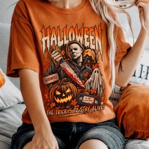 May include: An orange t-shirt featuring a graphic print with the text HALLOWEEN at the top and THE TRICK IS TO STAY ALIVE at the bottom. The design includes a masked figure holding a bloody knife, a jack-o'-lantern, a ghost, and a VHS tape.