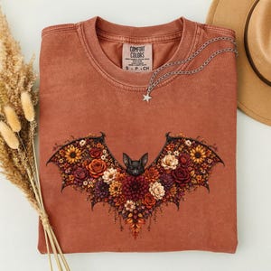May include: A terracotta-colored crewneck t-shirt featuring a graphic of a bat with wings composed of various flowers in shades of red, orange, and yellow. The shirt has a Comfort Colors label at the neck and a silver star pendant necklace.