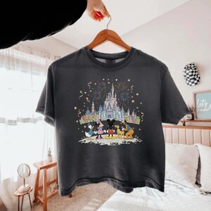 K&ouml;nnte beinhalten: Dunkelgraues T-Shirt mit einem farbenfrohen Aufdruck von Disney-Figuren vor einem Schloss mit Feuerwerk. Mickey, Minnie, Donald und Goofy sind abgebildet. Das T-Shirt h&auml;ngt an einem Holzb&uuml;gel.