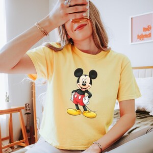 May include: A bright yellow short-sleeved t-shirt featuring a centered graphic of Mickey Mouse. The character is depicted with his signature black ears, red shorts with two white buttons, white gloves, and large yellow shoes, standing in a classic pose.