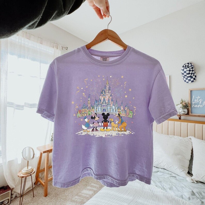 K&ouml;nnte beinhalten: Ein helllila T-Shirt mit einem Disney-Schloss- und Charakter-Grafik. Das Design zeigt Micky Maus, Minnie Maus, Donald Duck und Goofy. Das Shirt h&auml;ngt an einem Holzb&uuml;gel. Das Bild zeigt ein Feiertags-Thema.