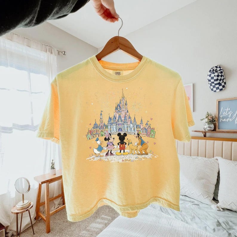 K&ouml;nnte beinhalten: Gelbes T-Shirt mit einem Disney-Schloss und Charakter-Grafik, darunter Micky und Minnie Maus, Donald Duck und Goofy. Das T-Shirt h&auml;ngt an einem Holzb&uuml;gel.