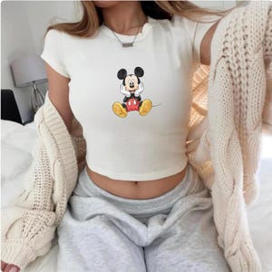May include: A cream-coloured, short-sleeved crop top with a Mickey Mouse graphic. The cartoon character has black ears, a black and red outfit, and yellow shoes. The top is worn with grey sweatpants and a cream-coloured cardigan.
