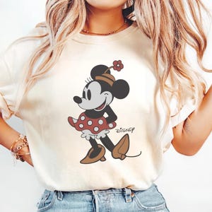 May include: Cream-colored t-shirt featuring a cartoon illustration of Minnie Mouse. Minnie is depicted in a red polka-dot dress, brown shoes, and a matching hat with a flower. The word "Disney" is printed below the character.
