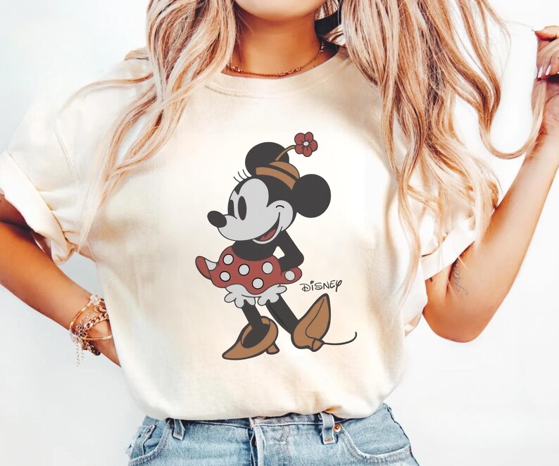 May include: Cream-colored t-shirt featuring a cartoon illustration of Minnie Mouse. Minnie is depicted in a red polka-dot dress, brown shoes, and a matching hat with a flower. The word "Disney" is printed below the character.
