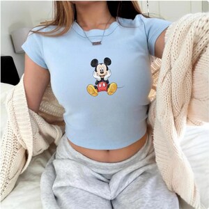 May include: Pale blue crop top with a Mickey Mouse design. The top has short sleeves and a round neck. The graphic shows Mickey Mouse with his hands on his face. A beige cardigan is draped over the shoulders.