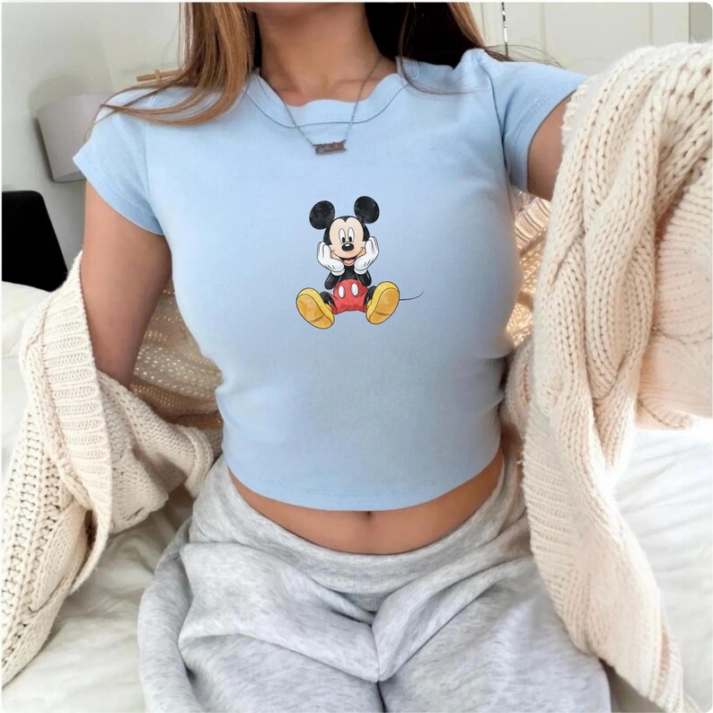 May include: Pale blue crop top with a Mickey Mouse design. The top has short sleeves and a round neck. The graphic shows Mickey Mouse with his hands on his face. A beige cardigan is draped over the shoulders.