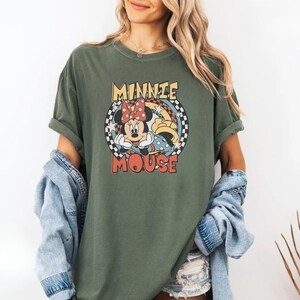 May include: An olive green t-shirt featuring a retro graphic of Minnie Mouse. The design includes Minnie's face, a red polka dot bow, and the words "MINNIE MOUSE" in a circular design with a checkered border. The shirt has rolled-up sleeves.