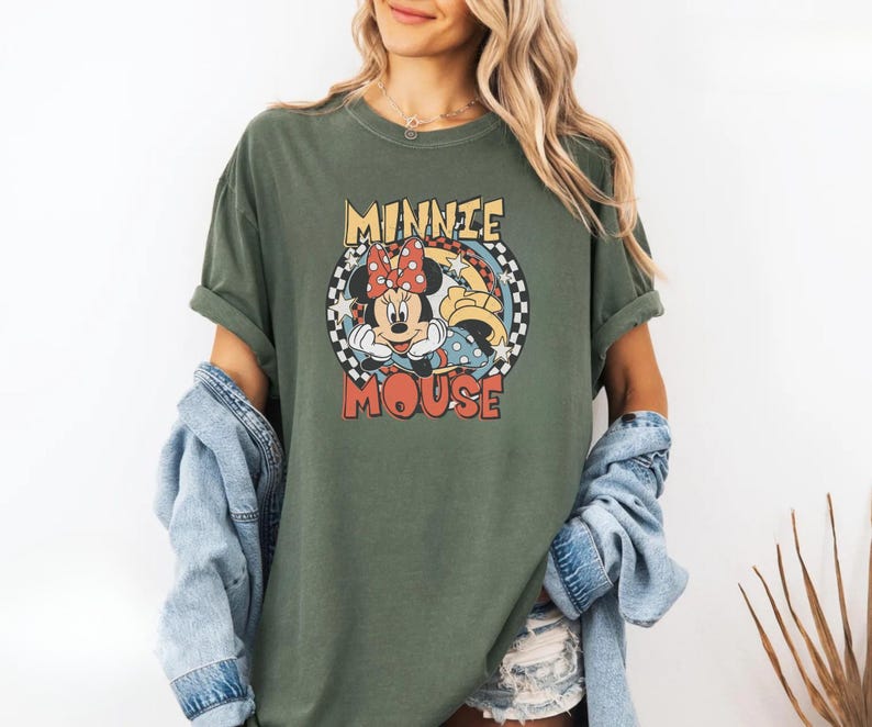 May include: An olive green t-shirt featuring a retro graphic of Minnie Mouse. The design includes Minnie's face, a red polka dot bow, and the words "MINNIE MOUSE" in a circular design with a checkered border. The shirt has rolled-up sleeves.