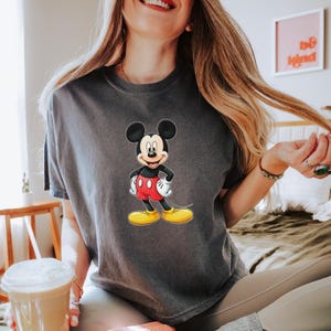 May include: A charcoal gray short-sleeve t-shirt featuring a centered graphic of Mickey Mouse. The character is depicted with his signature black ears, white gloves, red shorts, and yellow shoes, standing with hands on his hips in a classic pose.