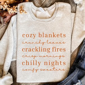 May include: A light gray crewneck sweatshirt featuring orange text that reads: cozy blankets, crunchy leaves, crackling fires, crisp mornings, chilly nights, comfy sweaters. The garment has long sleeves and a ribbed collar with a white interior tag.