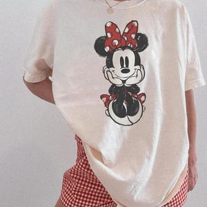 May include: Cream-coloured t-shirt featuring a Minnie Mouse graphic with a red bow and polka dots. The shirt is paired with red and white checked shorts. The overall style is casual and comfortable.