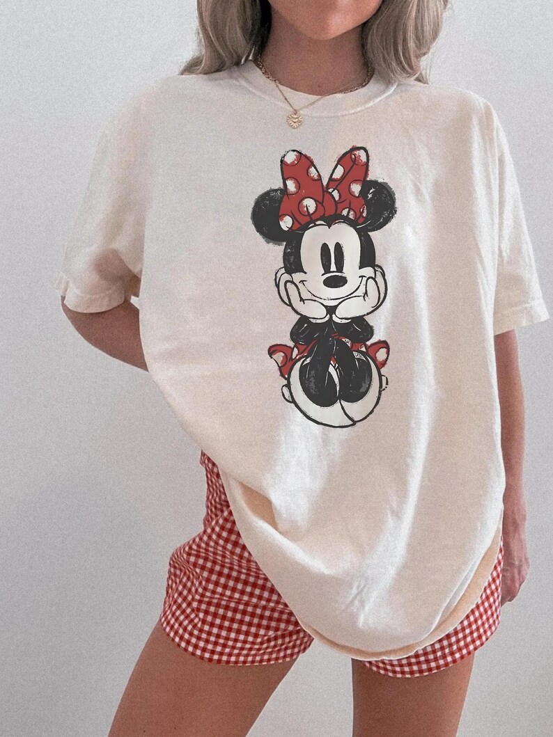 May include: Cream-coloured t-shirt featuring a Minnie Mouse graphic with a red bow and polka dots. The shirt is paired with red and white checked shorts. The overall style is casual and comfortable.
