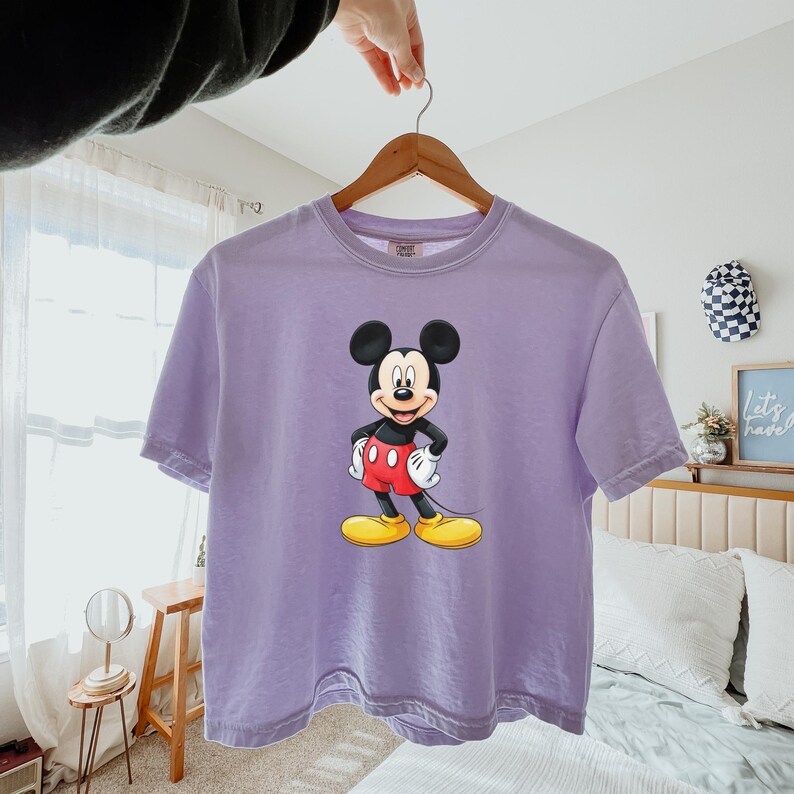 May include: A light purple short-sleeved t-shirt featuring a centered graphic of Mickey Mouse. The character is depicted with his signature black ears, red shorts, white gloves, and yellow shoes, standing with hands on his hips against the fabric.