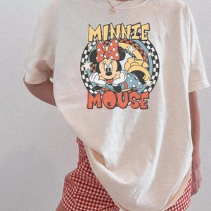 May include: Cream-colored t-shirt featuring a graphic of Minnie Mouse with the text "MINNIE MOUSE" in a retro style. The design includes a red bow, polka dots, and a checkered background. Paired with red and white checkered shorts.