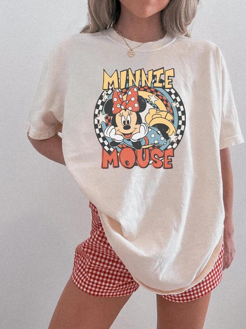 May include: Cream-colored t-shirt featuring a graphic of Minnie Mouse with the text "MINNIE MOUSE" in a retro style. The design includes a red bow, polka dots, and a checkered background. Paired with red and white checkered shorts.