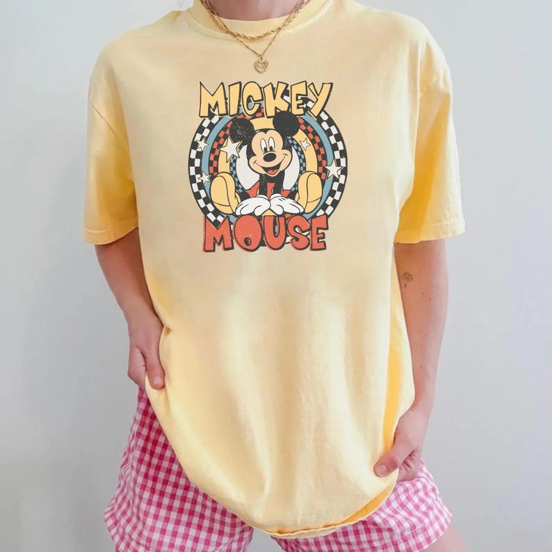 May include: Yellow t-shirt featuring a retro Mickey Mouse graphic. The design includes Mickey Mouse in a seated pose within a circular frame with the words "MICKEY" and "MOUSE". The shirt is a light yellow color.